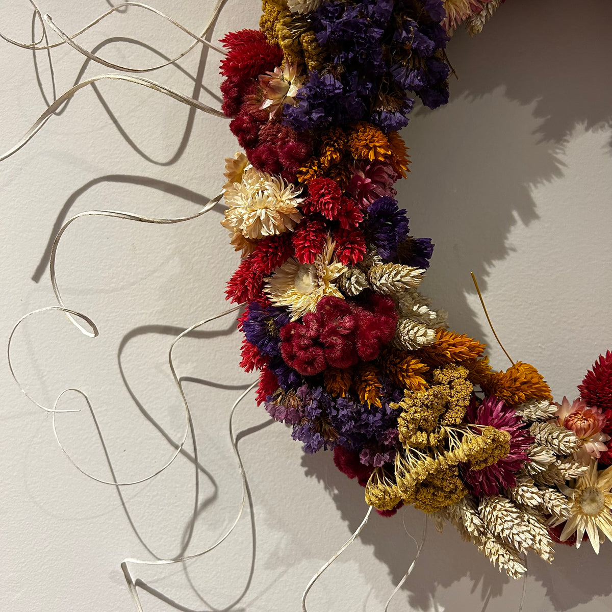 A dried flower wreath with a variety of colors including red, purple, yellow, and cream, hanging on a light-colored wall with visible shadows.