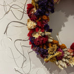 A dried flower wreath with a variety of colors including red, purple, yellow, and cream, hanging on a light-colored wall with visible shadows. 5