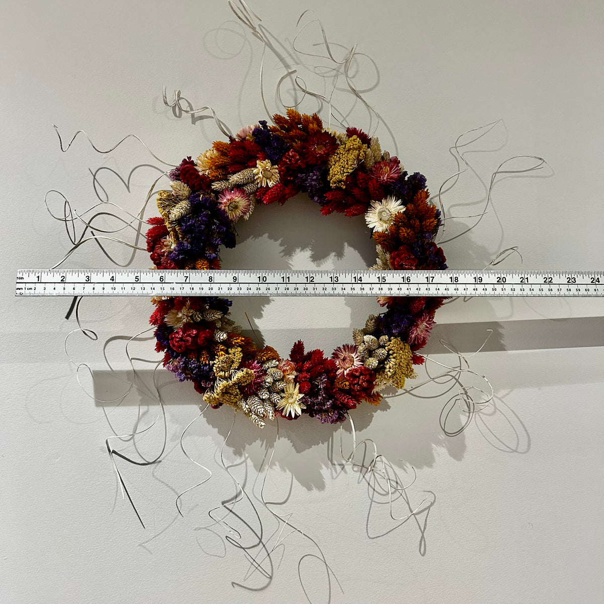 A circular dried flower wreath in autumnal reds, purples, cream, and gold tones, displayed against a white wall with a metal ruler horizontally aligned across the center for scale.