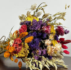 This magnificent, hand-tied bouquet is a vibrant symphony of contrasting colours and textures designed for the sophisticated interior. The Amethyst base of lush purple Statice provides a dramatic backdrop for the Gold accents: bright yellow Achillea Parker and soft natural pink Helichrysums. Textural elements featuring the unique, wavy shapes of red coral Celosia and Phalaris dyed in three complementary colors (natural, orange, and red).