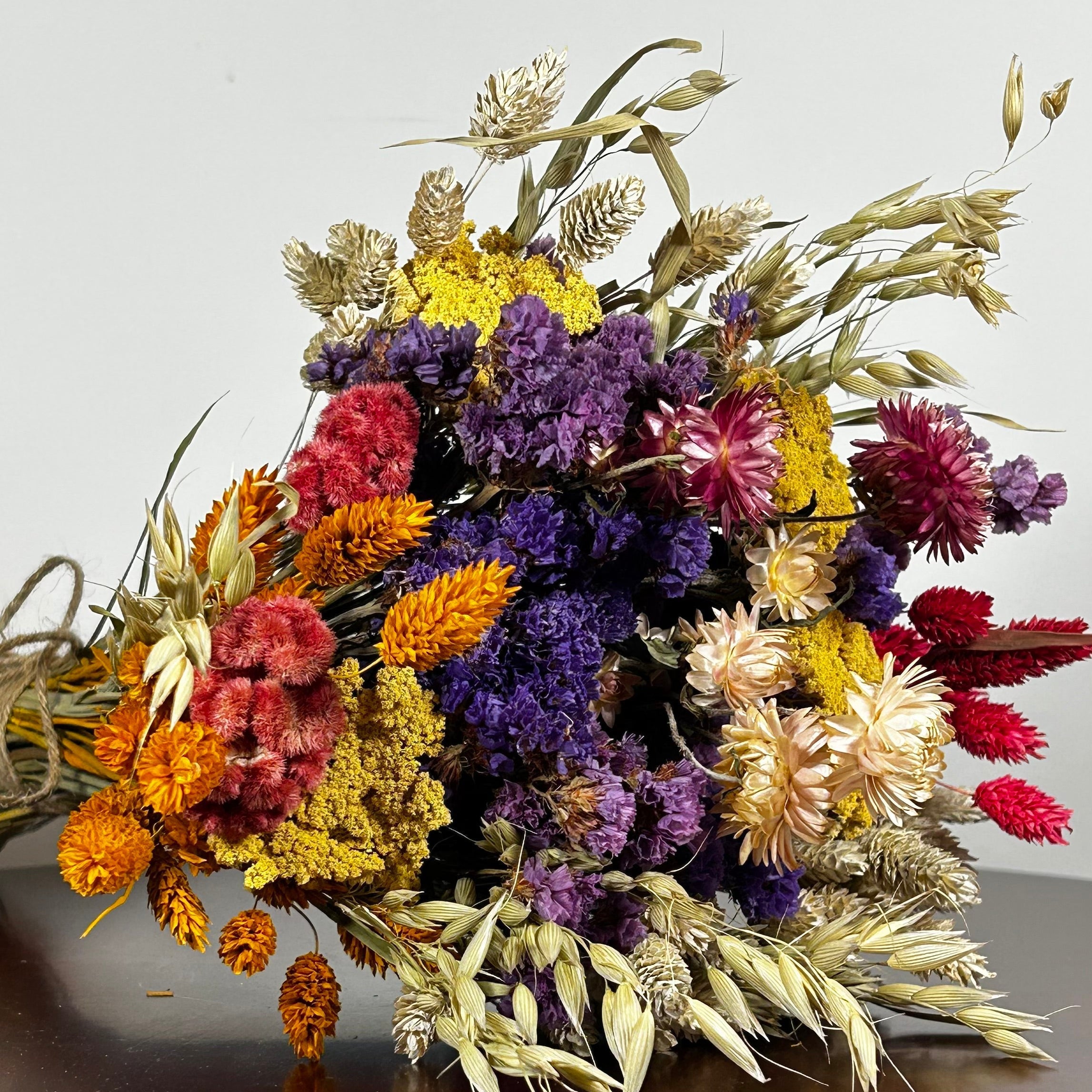 This magnificent, hand-tied bouquet is a vibrant symphony of contrasting colours and textures designed for the sophisticated interior. The Amethyst base of lush purple Statice provides a dramatic backdrop for the Gold accents: bright yellow Achillea Parker and soft natural pink Helichrysums. Textural elements featuring the unique, wavy shapes of red coral Celosia and Phalaris dyed in three complementary colors (natural, orange, and red).
