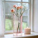 Blush coloured heart shaped glass vase with flowers and a candle on a windowsill.
