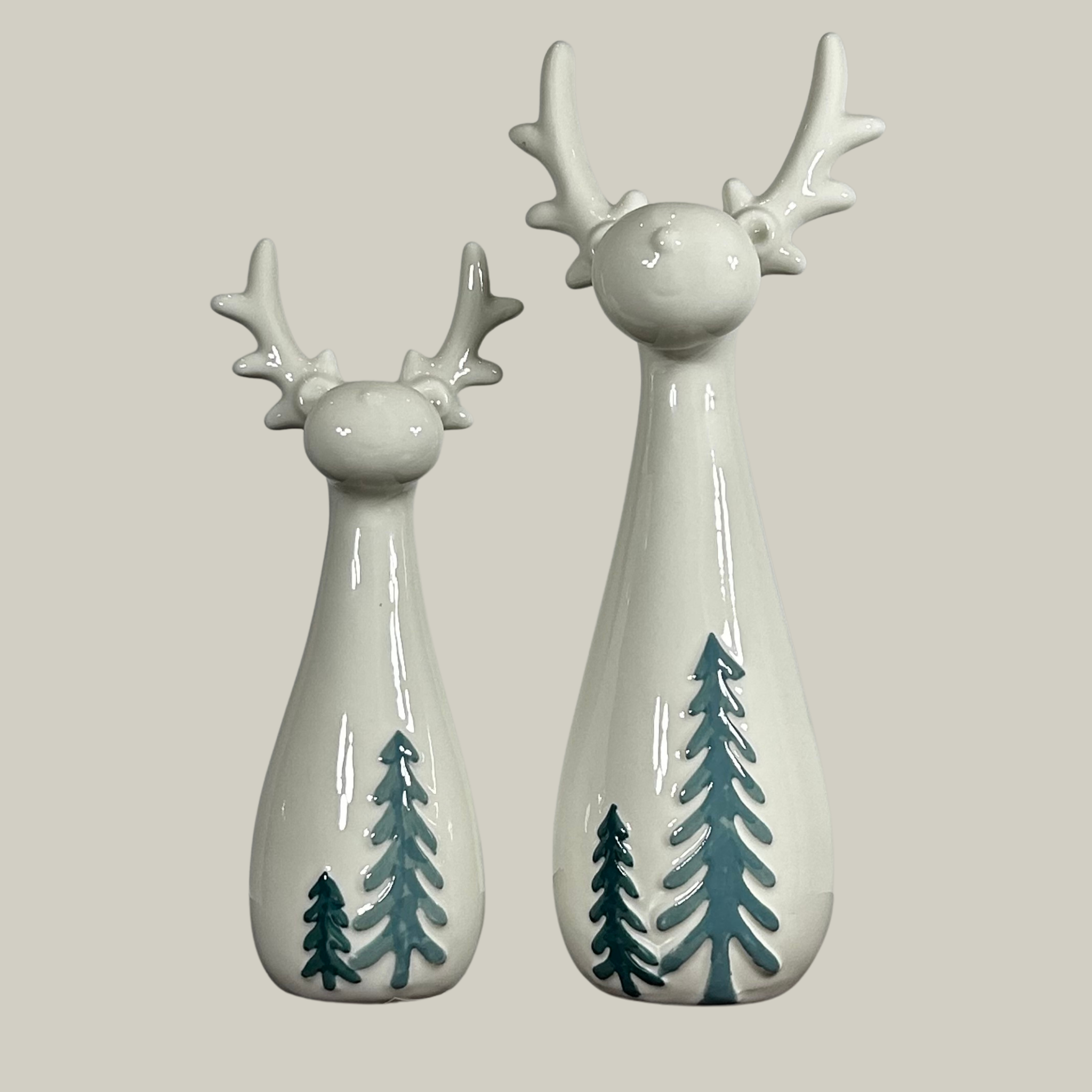 Two ceramic deer figurines in an ivory teardrop shape featuring the elegant silhouette of a deer's head and antlers. Embrace the whimsical detail of the miniature evergreen trees painted at the base, bringing a touch of nature's simple charm to the design.