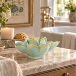 A light aqua and pale yellow ceramic water lily sculpture sits on a marble bathroom vanity next to a lit candle and flowers, with a bathtub and gold faucet in the softly blurred background.