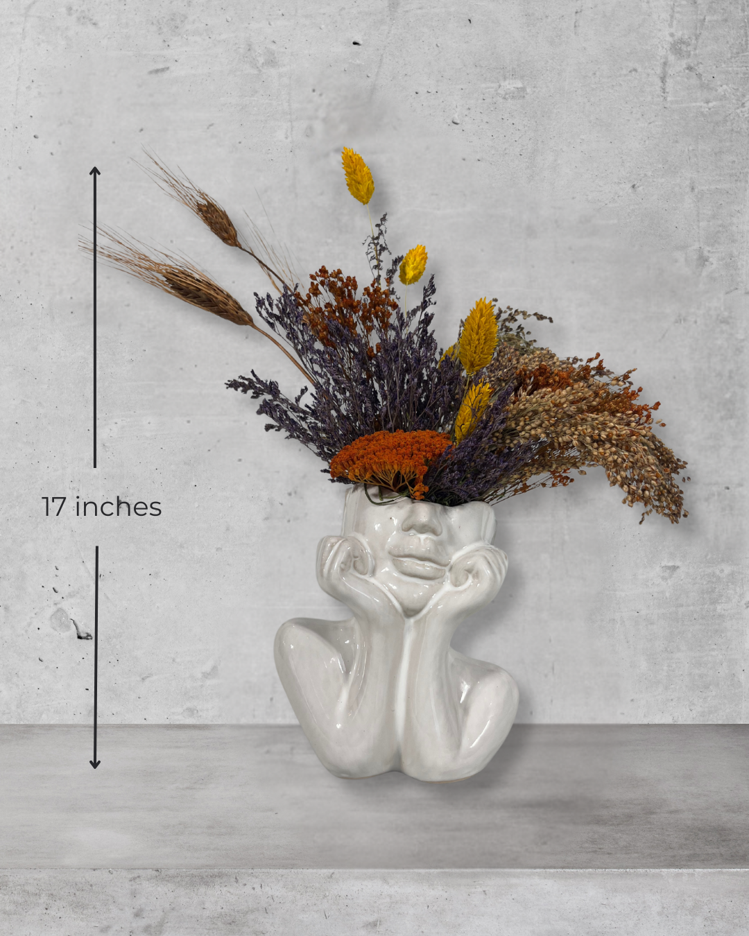White ceramic vase shaped like a face with dried flowers on a grey concrete background with measurement of 17 inches high