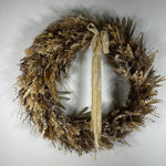 A rustic dried floral wreath in natural tan, brown, and beige tones, featuring a long cream ribbon bow hanging from the top center, photographed against a plain light gray background.