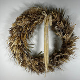 A rustic dried floral wreath in natural tan, brown, and beige tones, featuring a long cream ribbon bow hanging from the top center, photographed against a plain light gray background.