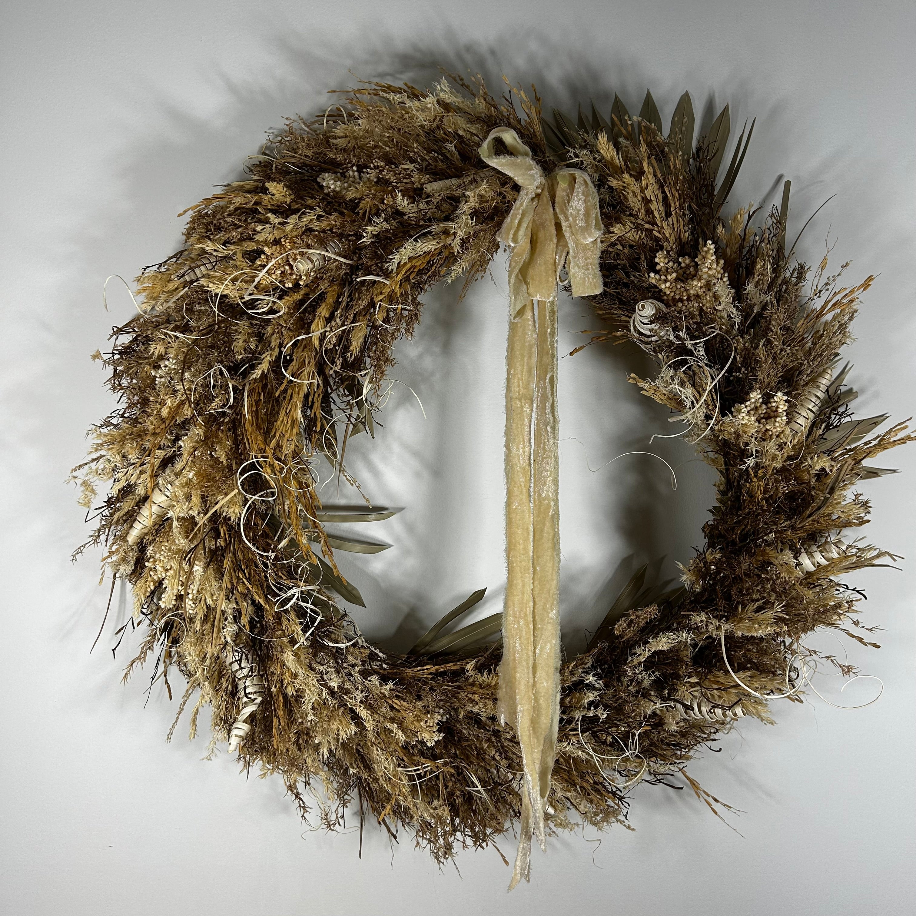 A rustic dried floral wreath in natural tan, brown, and beige tones, featuring a long cream ribbon bow hanging from the top center, photographed against a plain light gray background.
