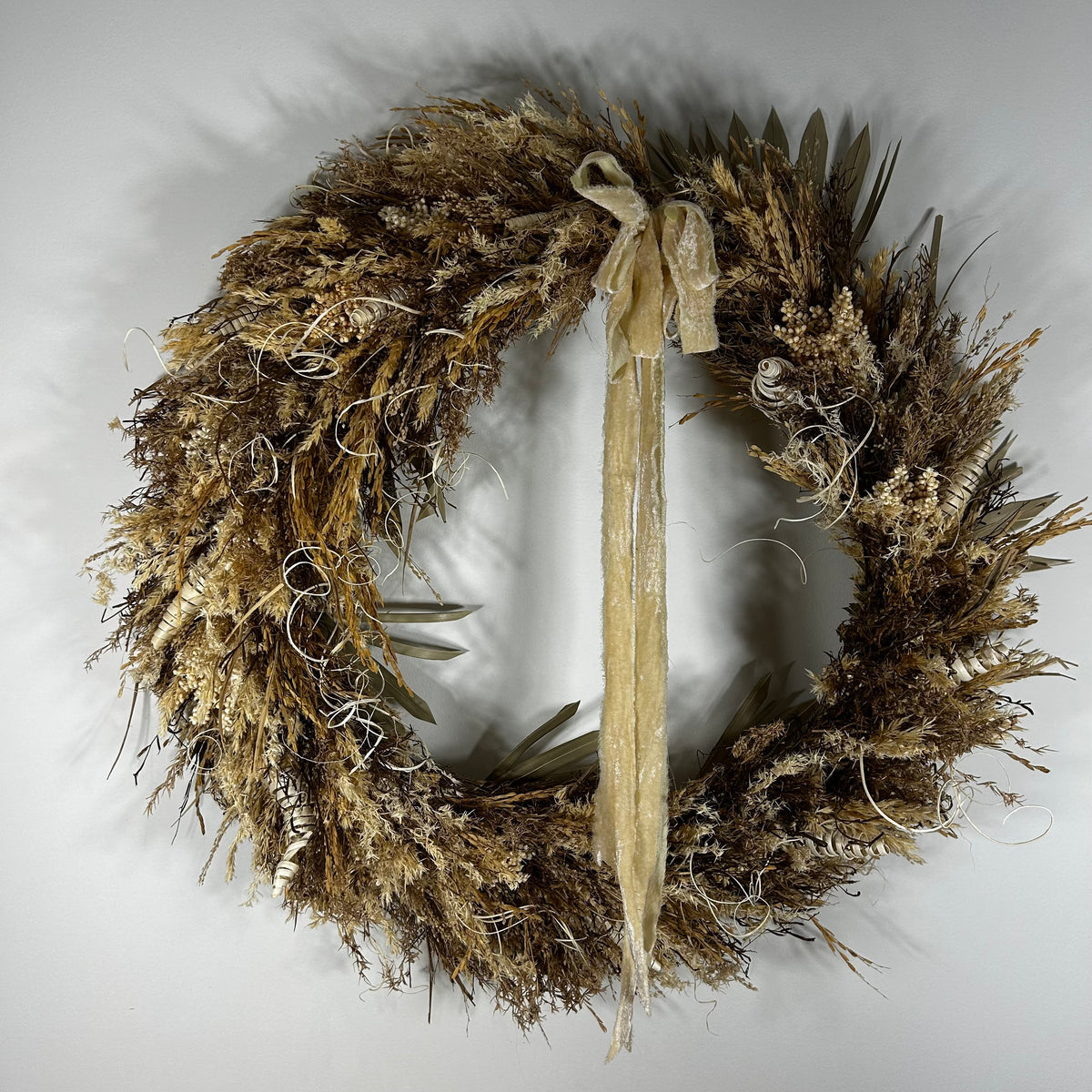 A rustic dried floral wreath in natural tan, brown, and beige tones, featuring a long cream ribbon bow hanging from the top center, photographed against a plain light gray background.