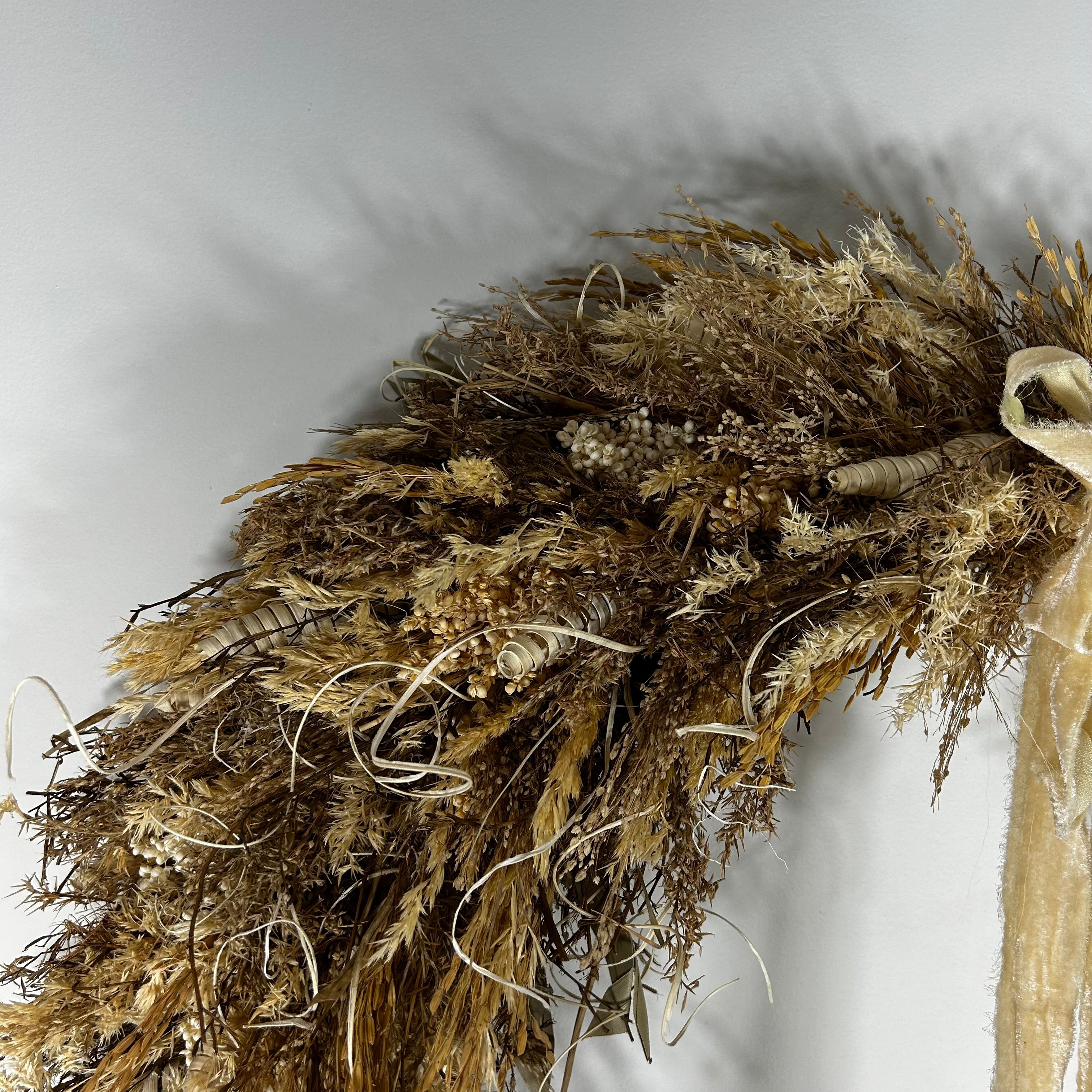 A dried floral arrangement featuring pampas grass, small beige berries, and a light tan ribbon, photographed against a plain light gray background.