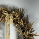 A dried floral wreath in natural beige, brown, and cream tones, featuring a large cream velvet bow, palm leaves, small dried flowers, and a seashell accent, photographed against a plain light gray wall.