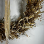 A dried botanical wall hanging featuring natural straw, reed, and moss textures in earthy tan, brown, and beige tones against a plain light background.
