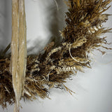 A dried botanical wall hanging featuring natural straw, reed, and moss textures in earthy tan, brown, and beige tones against a plain light background.