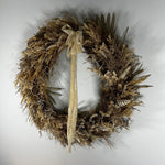 A rustic dried floral wreath made of natural brown and beige grasses, with a long beige ribbon bow hanging from the center, photographed against a plain light gray background.