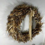 A rustic dried floral wreath in natural tan and brown tones, decorated with a long cream ribbon bow and small seashell accents, hanging against a plain white wall.