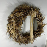A rustic dried floral wreath in natural tan and brown tones, decorated with a long cream ribbon bow and small seashell accents, hanging against a plain white wall.