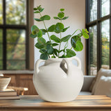 White ceramic vase with green plant on a wooden table in a room with large windows.