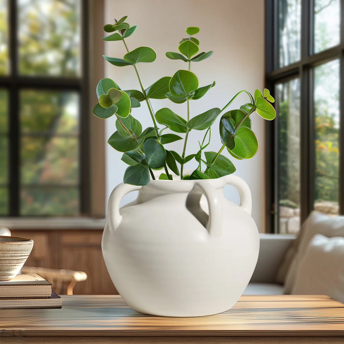 White ceramic vase with green plant on a wooden table in a room with large windows.