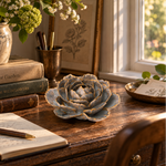 A decorative ceramic rose sculpture in muted blue-gray with coral toned edging sits on a rustic wooden desk in a sunlit room. Books, a vase of flowers, a pen, and framed artwork are visible around it.