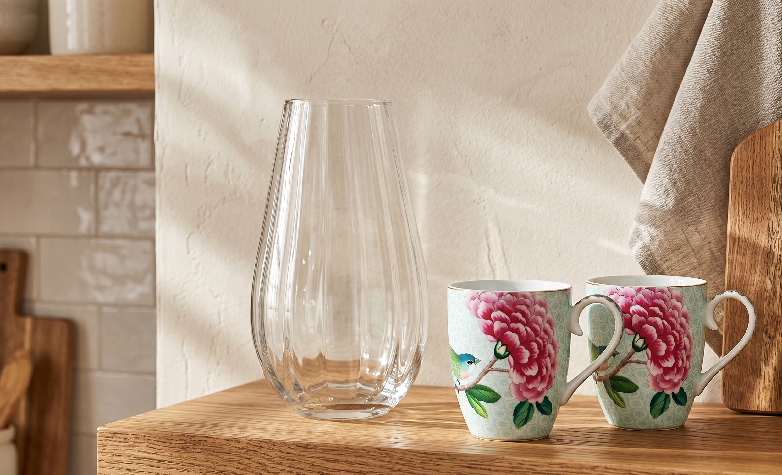 Two floral mugs and a clear vase on a wooden shelf in a kitchen setting.