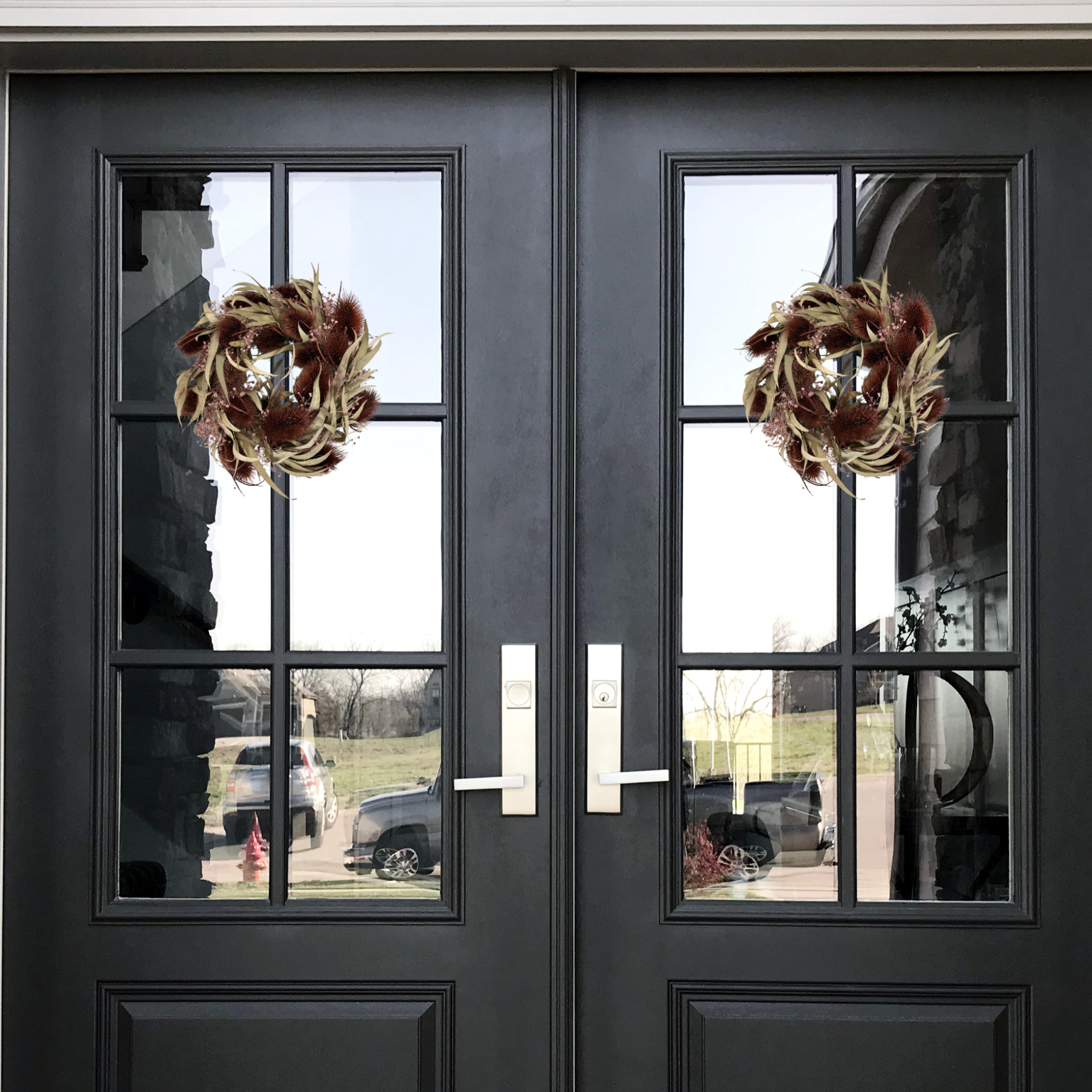 Black double doors with glass panels featuring two meticulously crafted 10-inch accent wreath with it's lush burgundy red teasels providing a striking focal point against the delicate, ethereal sprays of Bupleurum in a soft pink and natural willow eucalyptus adds a graceful touch of olive green.