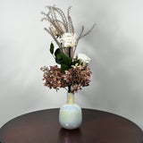 An preserved floral arrangement featuring white roses, dusty mauve hydrangeas, and tall textured beige stems, displayed in a glossy iridescent white vase on a dark brown wooden table against a light gray wall.