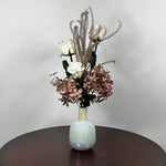 An preserved floral arrangement featuring white roses, dusty mauve hydrangeas, and tall textured beige stems, displayed in a glossy iridescent white vase on a dark brown wooden table against a light gray wall.