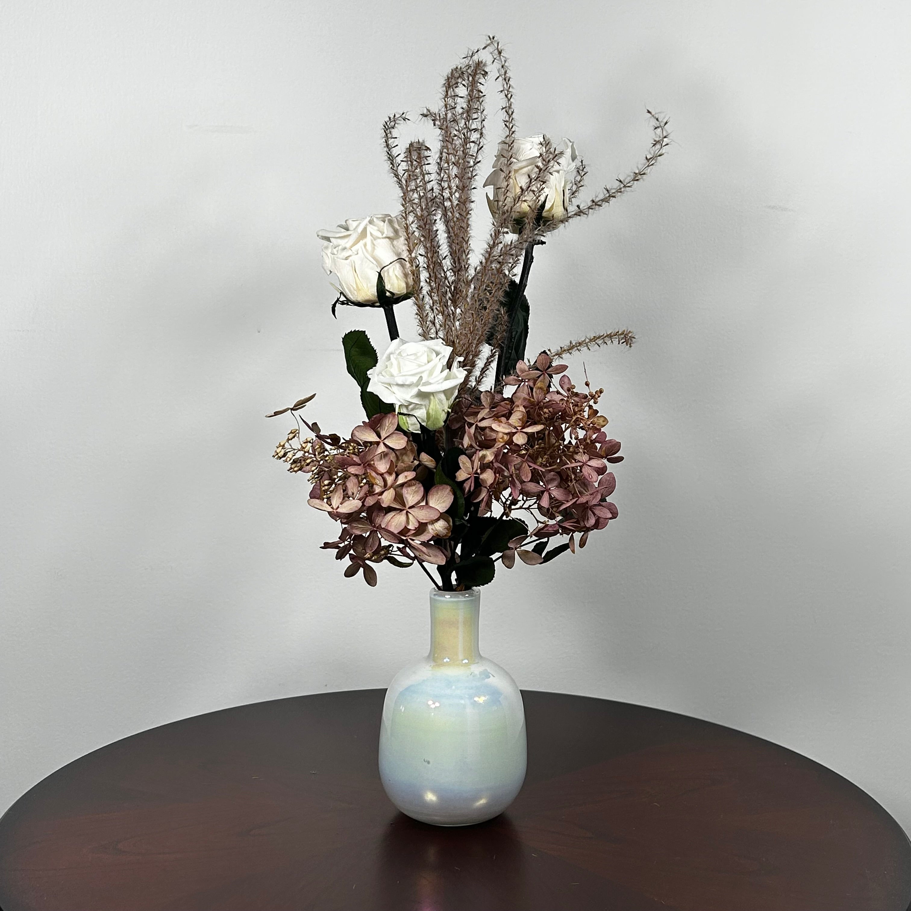 An preserved floral arrangement featuring white roses, dusty mauve hydrangeas, and tall textured beige stems, displayed in a glossy iridescent white vase on a dark brown wooden table against a light gray wall.