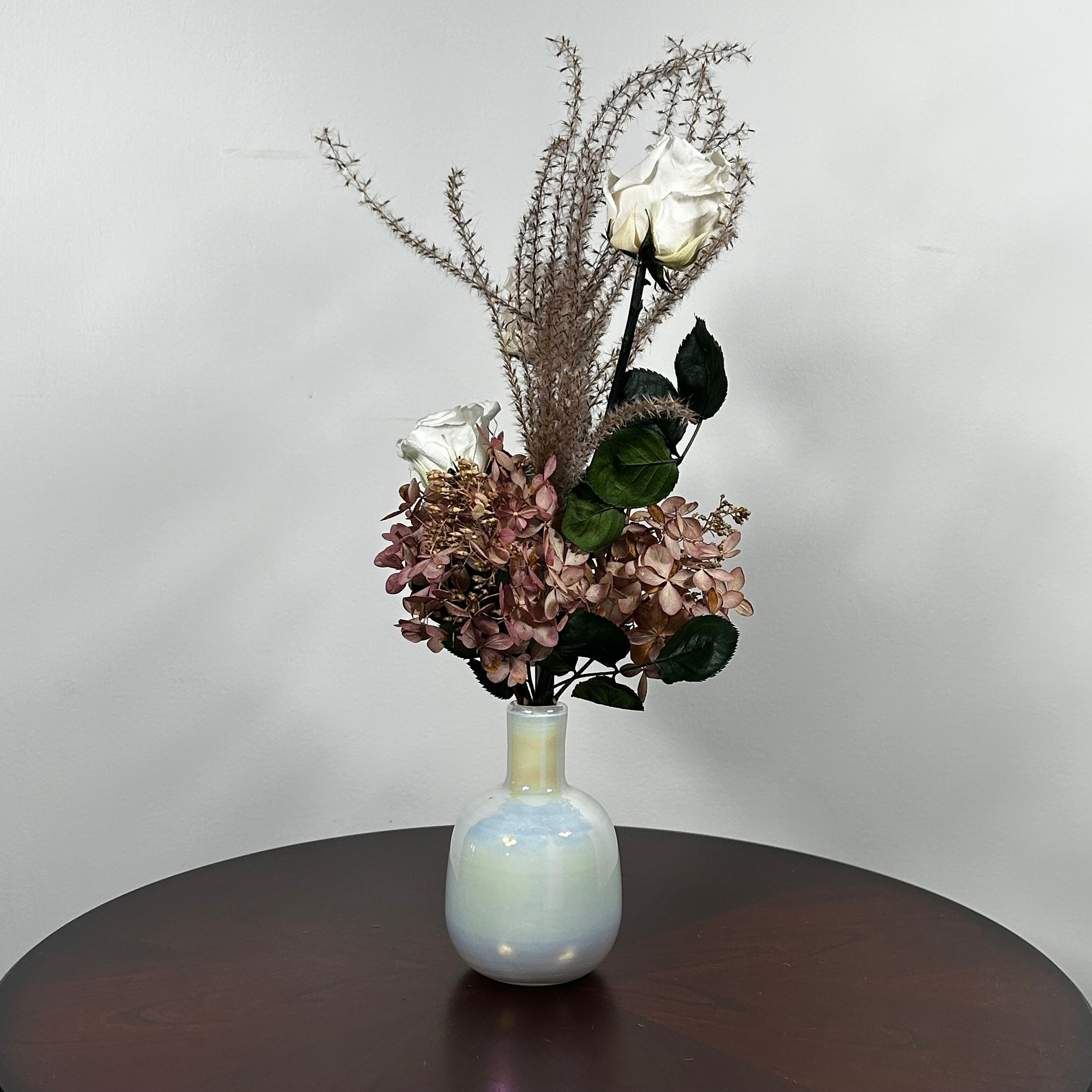 An preserved floral arrangement featuring white roses, dusty mauve hydrangeas, and tall textured beige stems, displayed in a glossy iridescent white vase on a dark brown wooden table against a light gray wall.