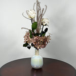 An preserved floral arrangement featuring white roses, dusty mauve hydrangeas, and tall textured beige stems, displayed in a glossy iridescent white vase on a dark brown wooden table against a light gray wall.