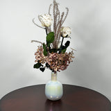 An preserved floral arrangement featuring white roses, dusty mauve hydrangeas, and tall textured beige stems, displayed in a glossy iridescent white vase on a dark brown wooden table against a light gray wall.