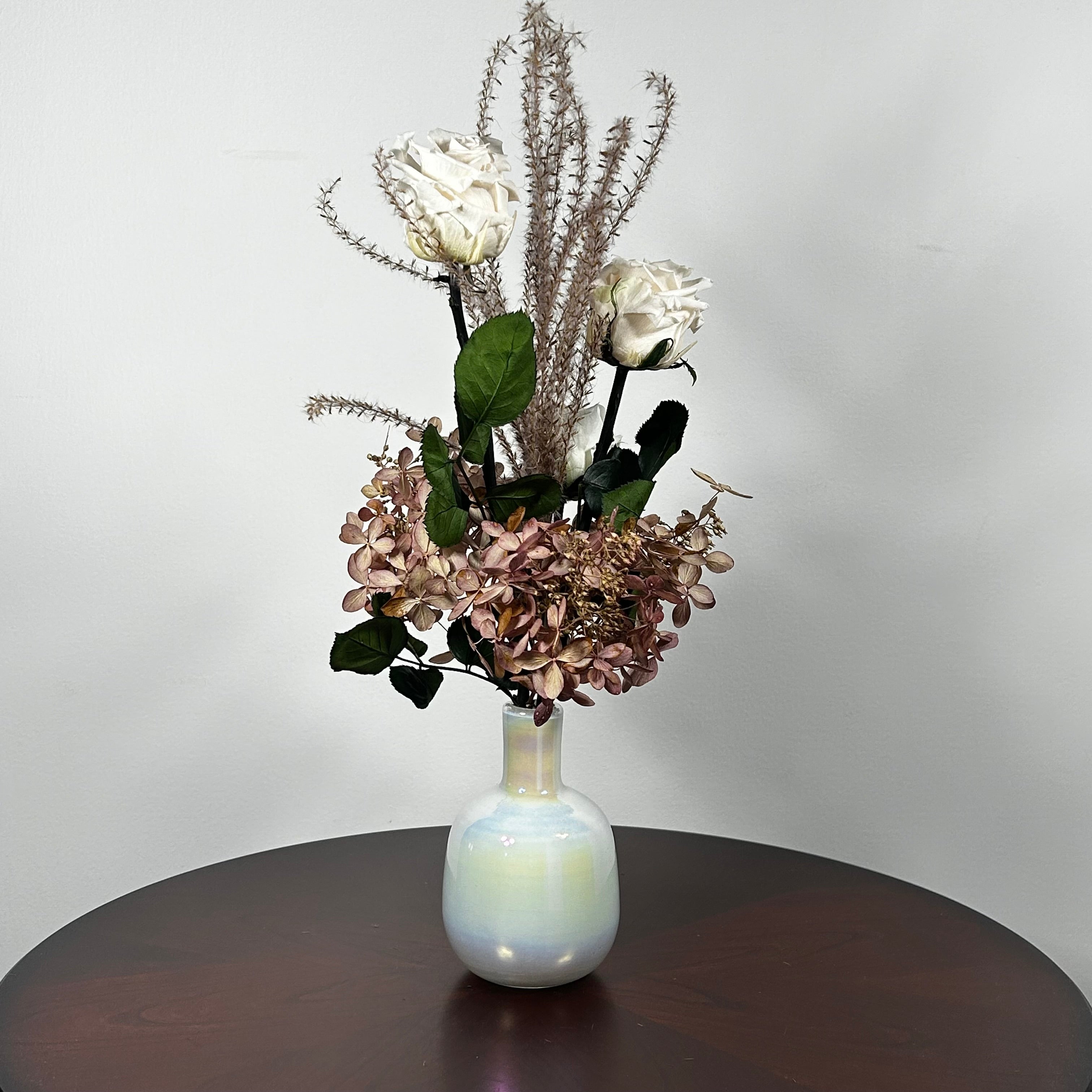 An preserved floral arrangement featuring white roses, dusty mauve hydrangeas, and tall textured beige stems, displayed in a glossy iridescent white vase on a dark brown wooden table against a light gray wall.