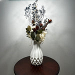 A white textured ceramic vase filled with a dried floral arrangement, placed on a round dark wood table against a plain light gray wall.