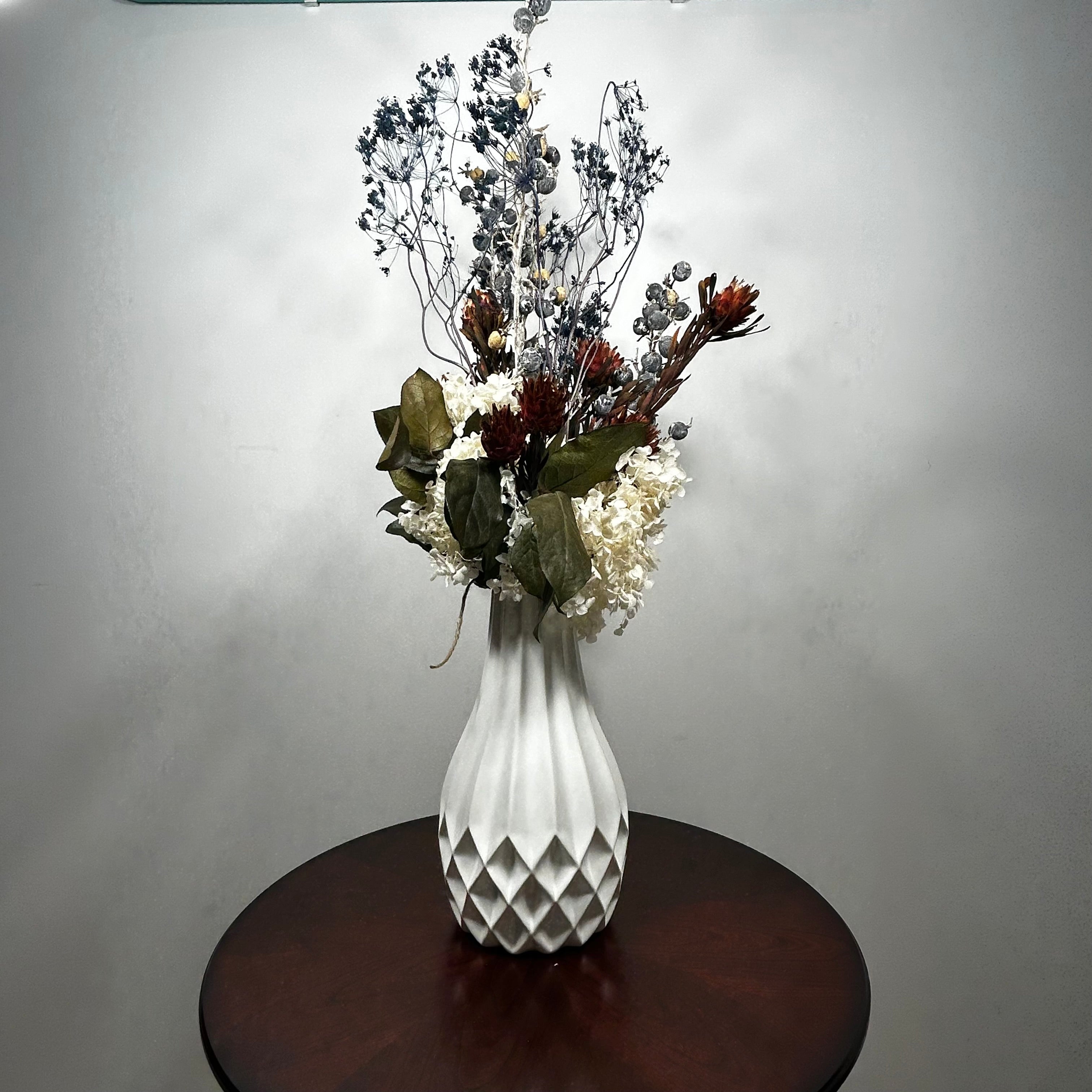 A white textured ceramic vase filled with a dried floral arrangement, placed on a round dark wood table against a plain light gray wall.