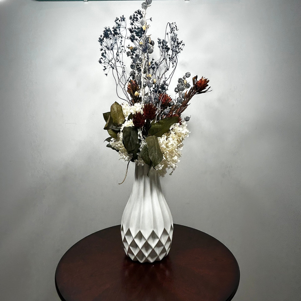 A white textured ceramic vase filled with a dried floral arrangement, placed on a round dark wood table against a plain light gray wall.