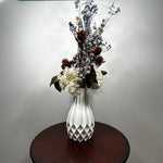 A white textured ceramic vase filled with a dried floral arrangement featuring white, burgundy, and gray botanicals, displayed on a round dark wood table against a plain light gray wall.