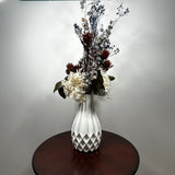 A white textured ceramic vase filled with a dried floral arrangement featuring white, burgundy, and gray botanicals, displayed on a round dark wood table against a plain light gray wall.
