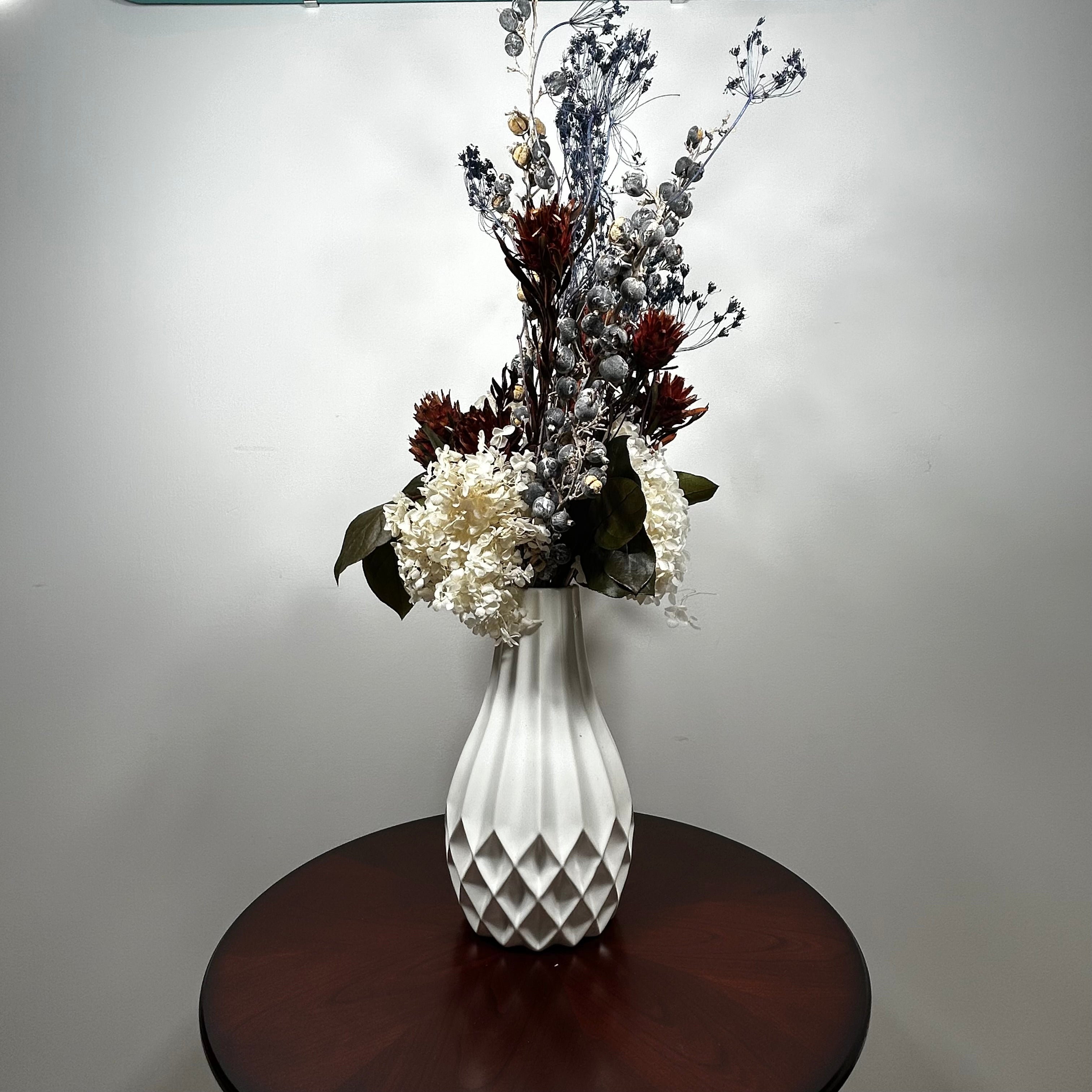 A white textured ceramic vase filled with a dried floral arrangement featuring white, burgundy, and gray botanicals, displayed on a round dark wood table against a plain light gray wall.