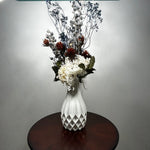 A white ceramic vase with a textured diamond pattern, filled with a dried floral arrangement featuring white, red, and blue flowers, set on a dark round wooden table against a neutral gray wall.