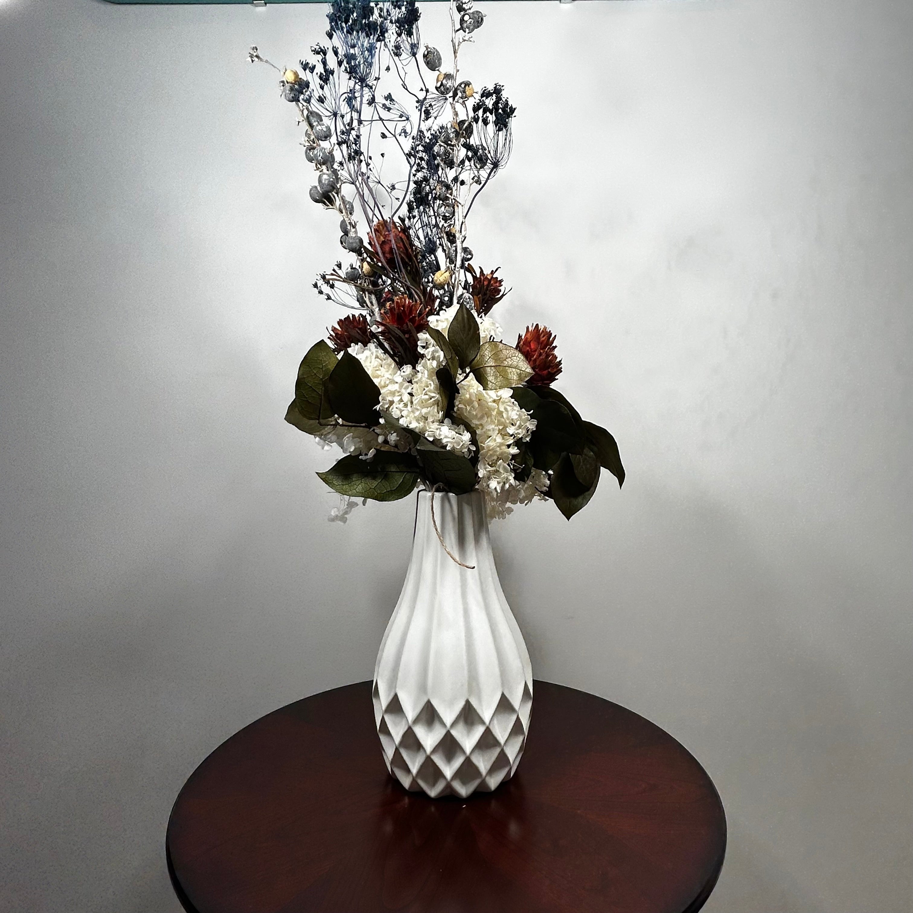 A white ribbed ceramic vase filled with a dried floral arrangement, displayed on a round dark wood table against a plain light gray wall.