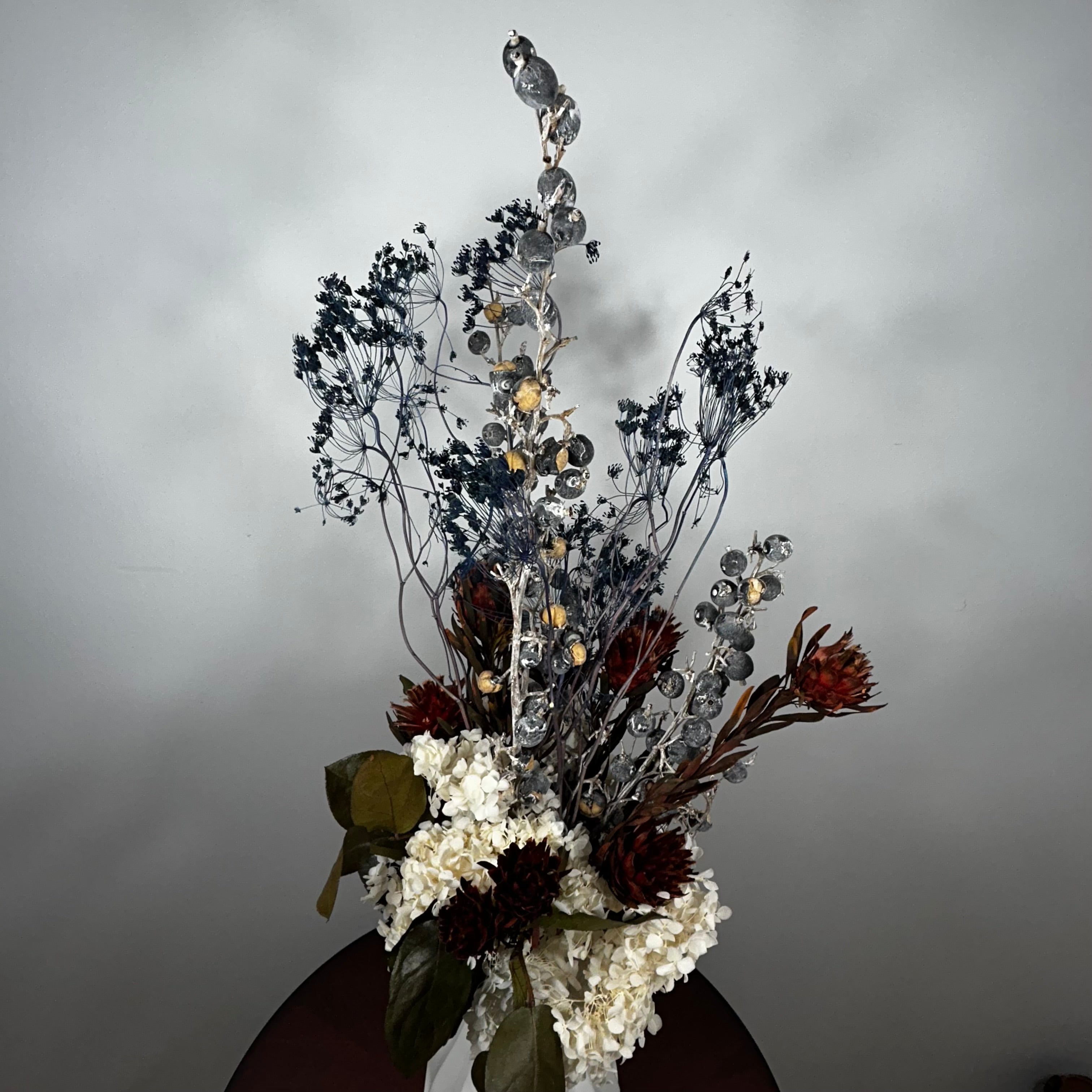 A dried floral arrangement featuring white hydrangeas, dark brown and rust protea flowers, and delicate blue-gray baby's breath on a silver branch, set against a plain gray wall.