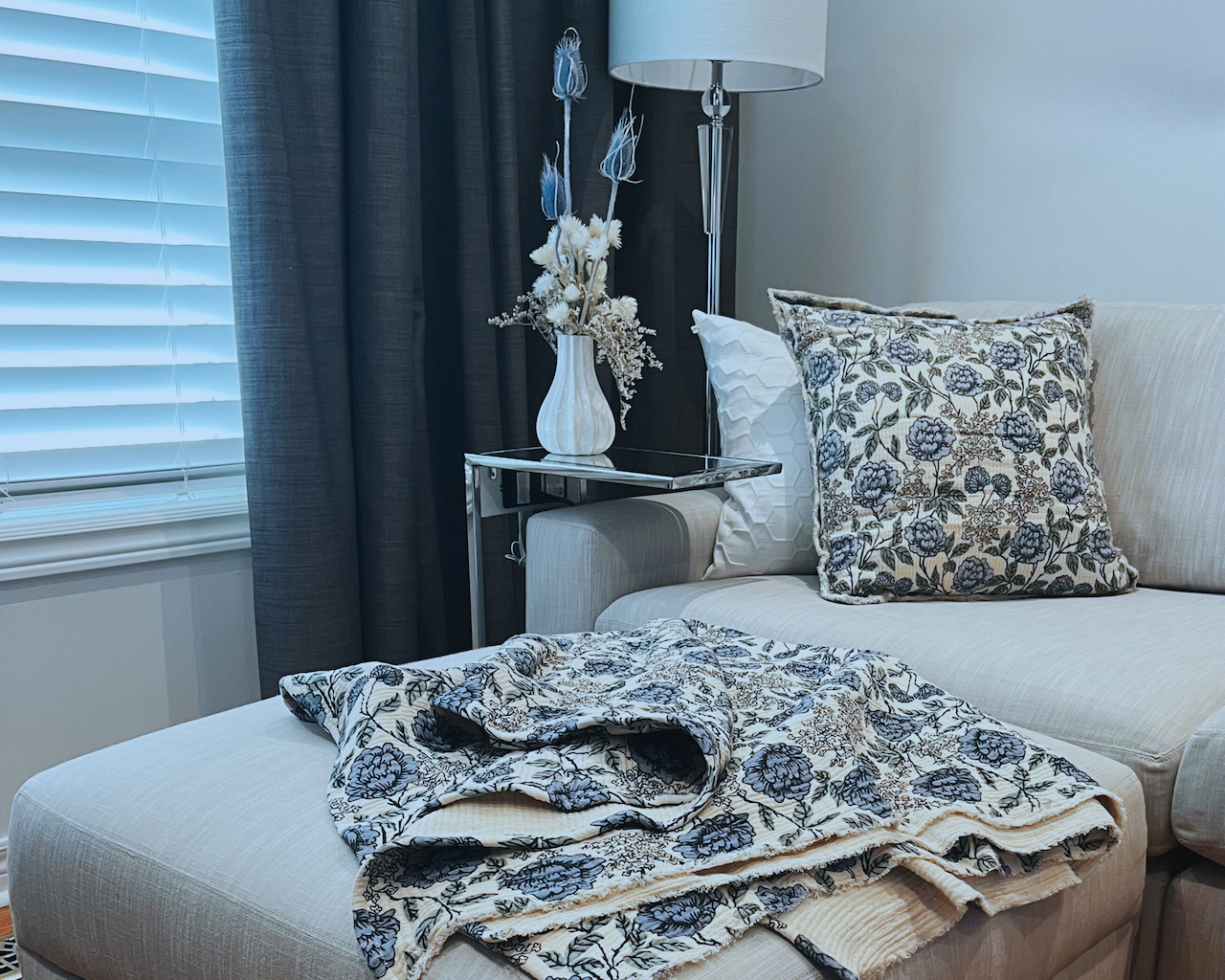 Living room with a soft grey sofa, blue and ivory floral-patterned cushions and matching throw, and a small table with a dried flower arrangement in a vase.