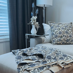 Living room with a soft grey sofa, blue and ivory floral-patterned cushions and matching throw, and a small table with a dried flower arrangement in a vase.