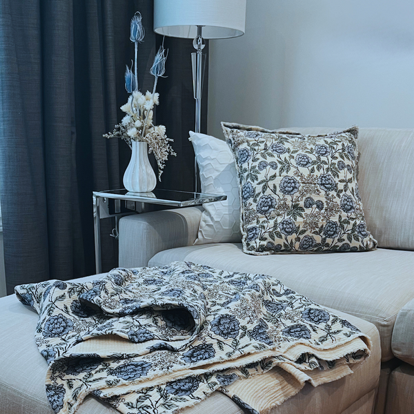 Living room with a soft grey sofa, blue and ivory floral-patterned cushions and matching throw, and a small table with a dried flower arrangement in a vase.