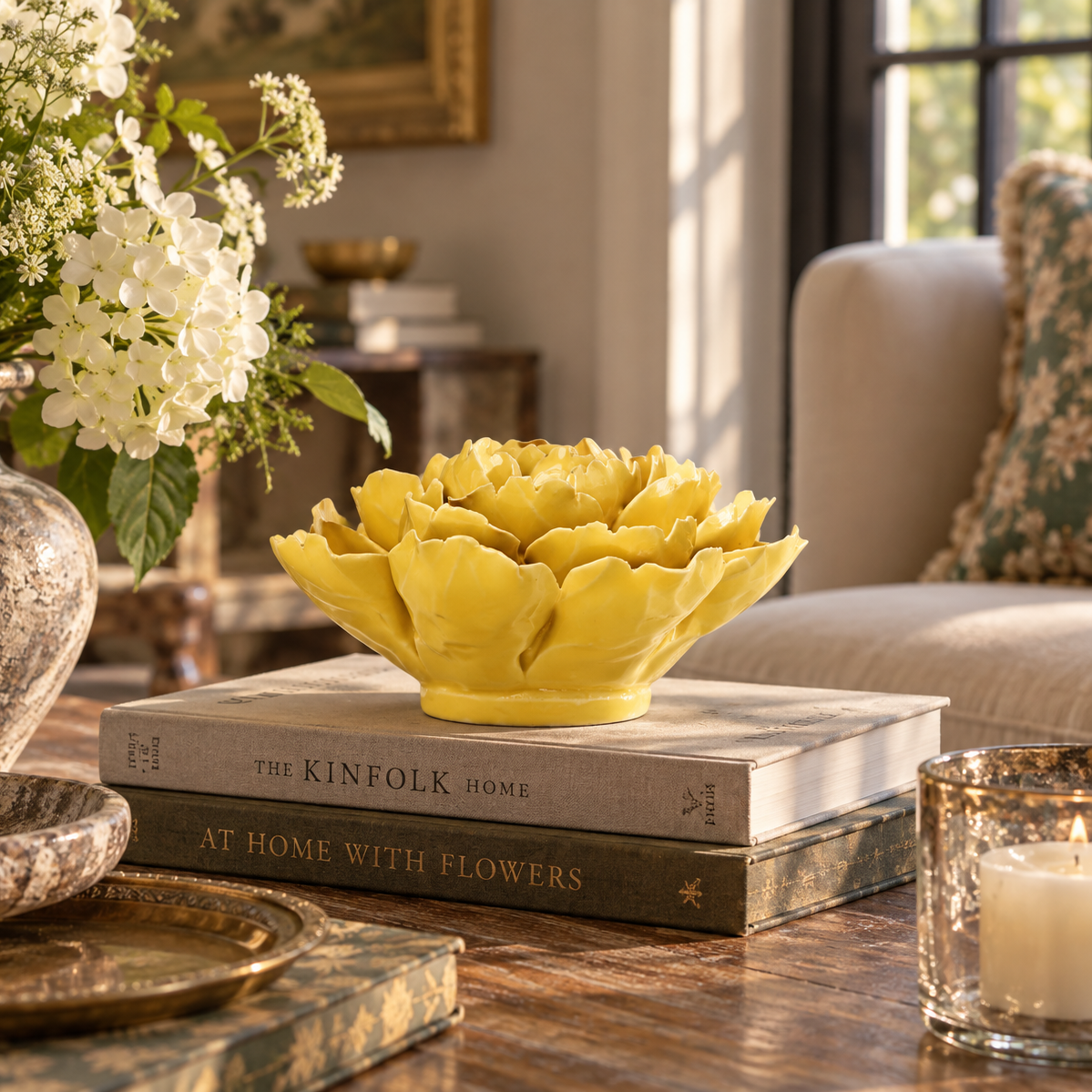 A bright yellow ceramic peony-shaped flower sits on top of two stacked books on a wooden table in a styled living room setting.