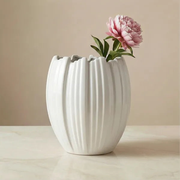A white ribbed ceramic vase with a flared rim, holding a single pink peony flower, set on a light marble surface against a warm beige background.