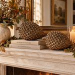 Two large metallic gold pinecones displayed on a fireplace mantel with stacked books, candles in glass holders, autumn leaves, and a vase of dried foliage in a warm, decorated interior setting.