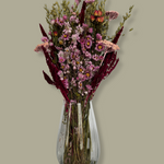 This striking, hand-tied bouquet is a harmonious blend of dried botanicals in deep red, soft pink, and natural oats. In this image the bouquet is in a clear glass vase against a plain green background.