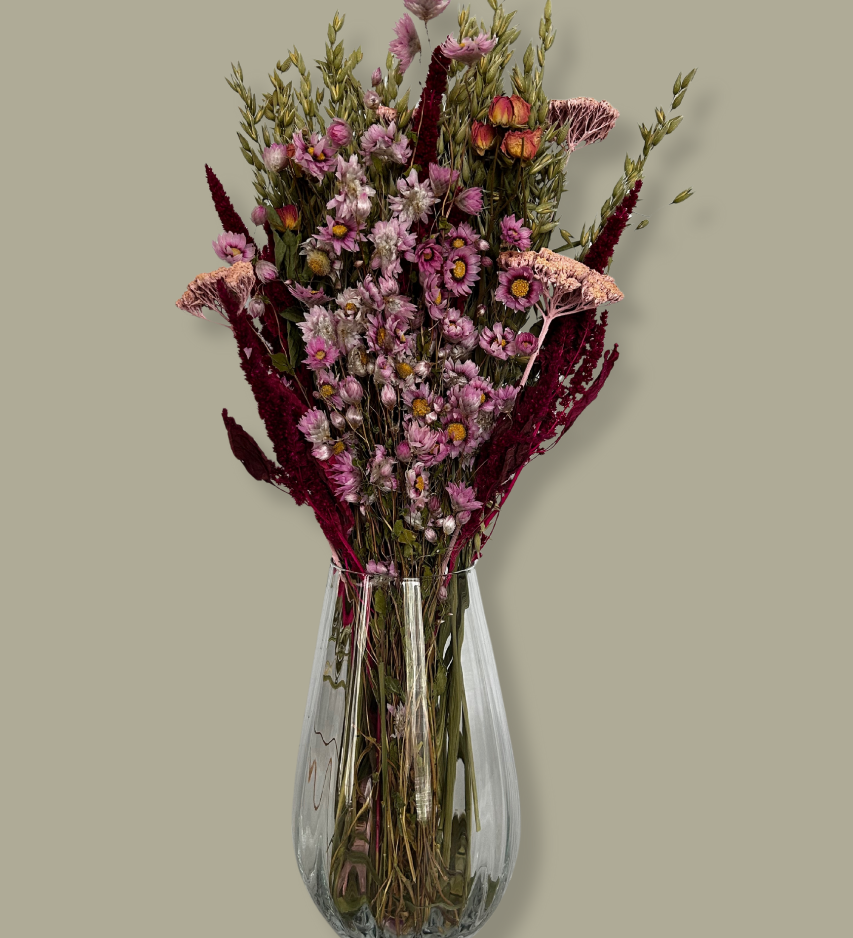This striking, hand-tied bouquet is a harmonious blend of dried botanicals in deep red, soft pink, and natural oats. In this image the bouquet is in a clear glass vase against a plain green background.