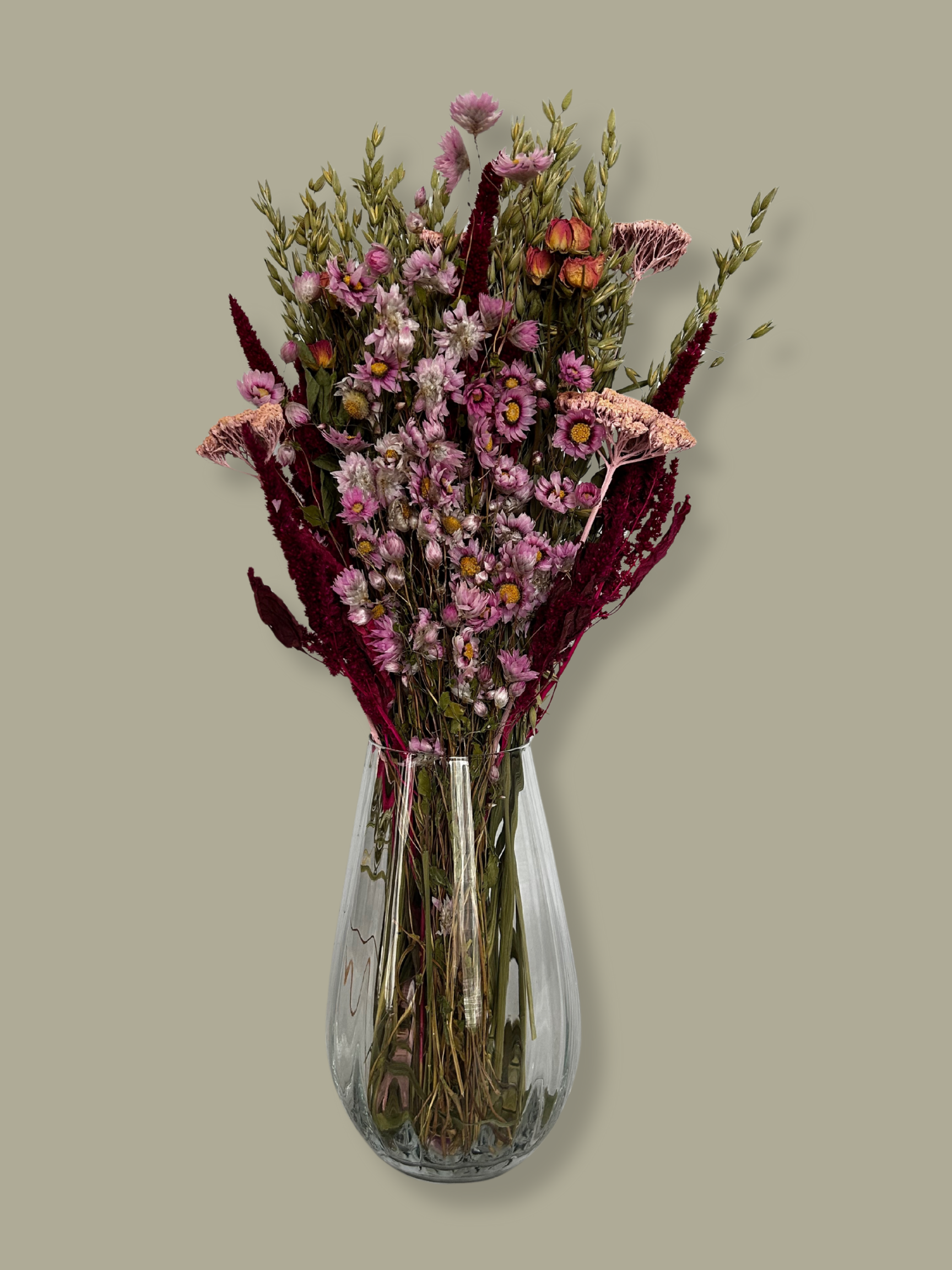 This striking, hand-tied bouquet is a harmonious blend of dried botanicals in deep red, soft pink, and natural oats. In this image the bouquet is in a clear glass vase against a plain green background.
